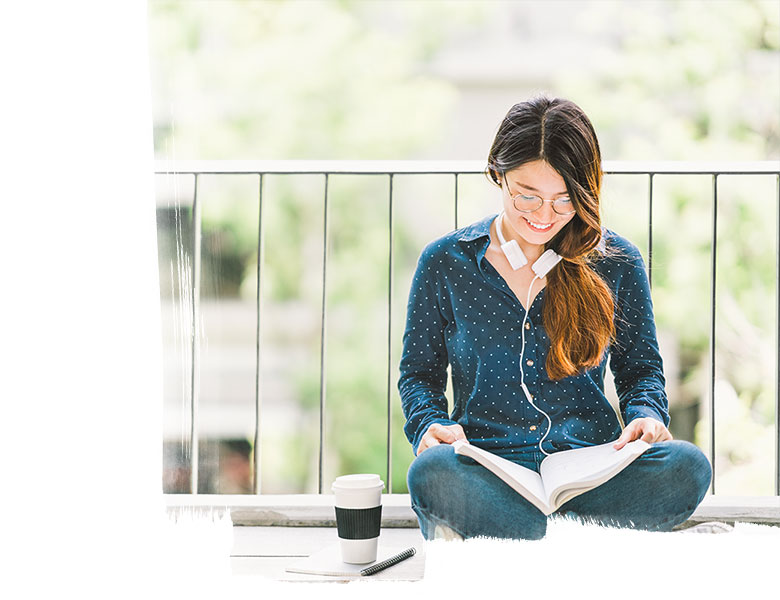A female student sitting cross-legged on the floor, smiling while reading a book (coffee cup beside her)