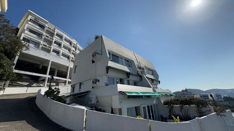 A multi-story white villa with green window awnings is situated on a steep, curved driveway.