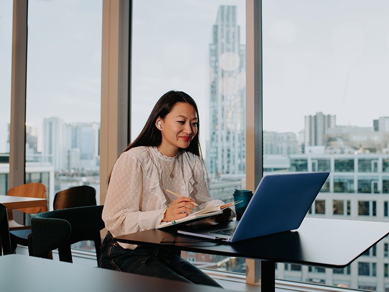 A content and confident young woman sits behind a laptop, taking notes.